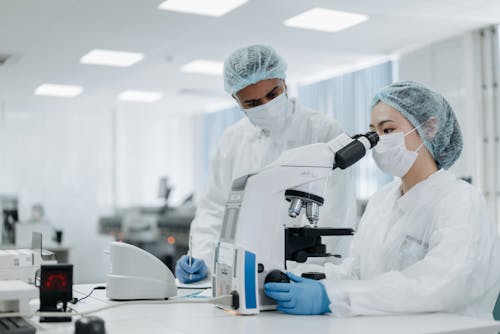 Free Woman in White Protective Suit Wearing  White Face Mask Looking Through the Microscope Stock Photo
