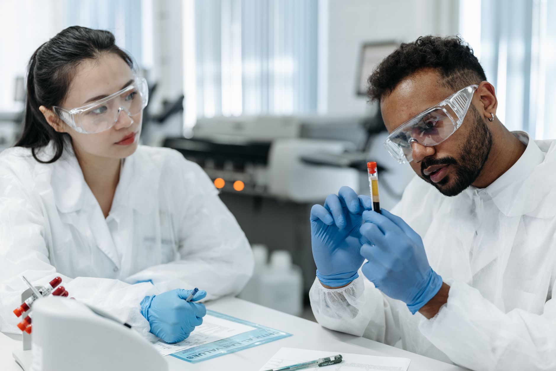 Two scientists in protective gear analyze blood samples in a laboratory setting.