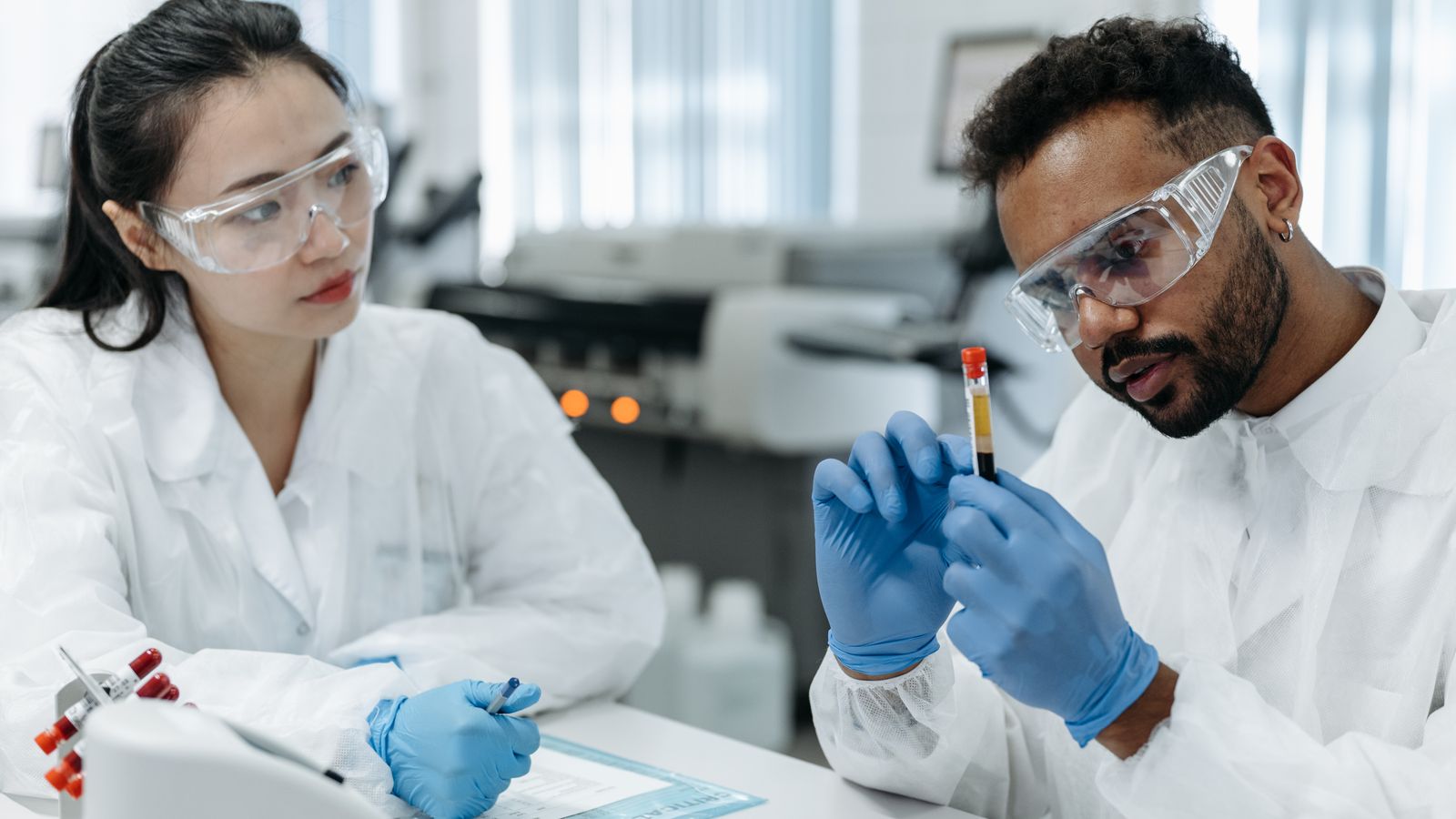 Two scientists in protective gear analyze blood samples in a laboratory setting.
