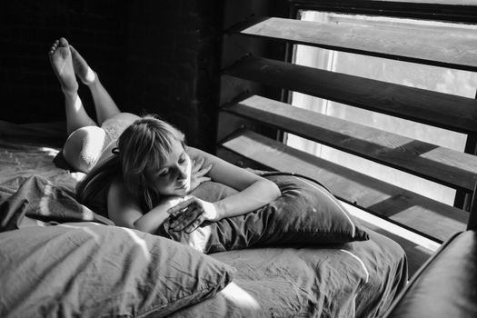 Black-and-white image of a woman lying on a bed by window, embracing tranquility.