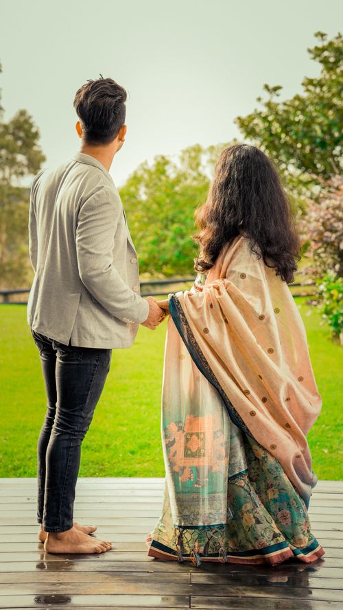 Free A couple stands holding hands on a wooden platform, wearing traditional attire, gazing into the lush scenery. Stock Photo