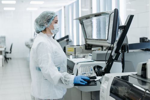 Free Woman in lab coat, face mask, and gloves working at a laboratory computer station. Stock Photo