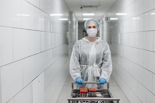 Healthcare worker in protective gear pushes cart with blood samples in sterile hospital corridor.
