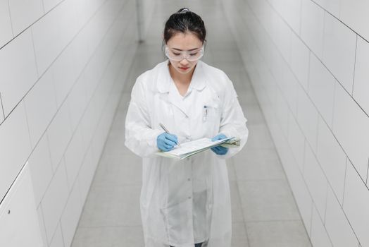 A female scientist wearing safety goggles and a lab coat taking notes in a laboratory corridor.