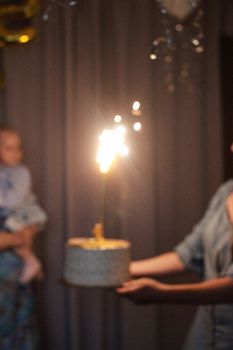 A sparklers-lit birthday cake being held at a blurry party.