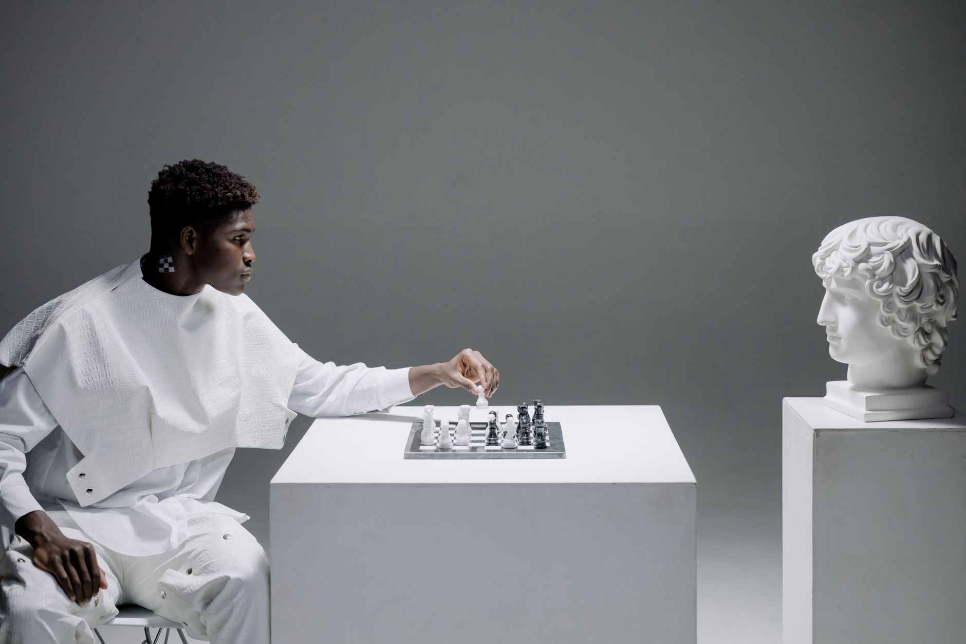 A man plays chess against a white sculpture bust, symbolizing strategy and thought.