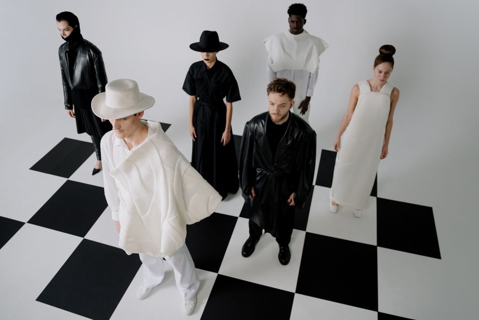 High-fashion models wear monochrome outfits on a chessboard pattern floor, showcasing diversity and style.