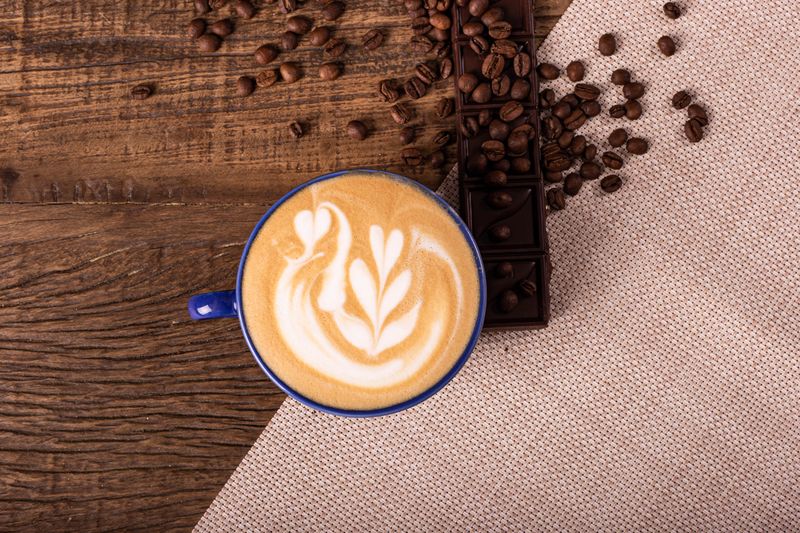 Overhead view of a latte with swan latte art, surrounded by coffee beans and chocolate on rustic wood.