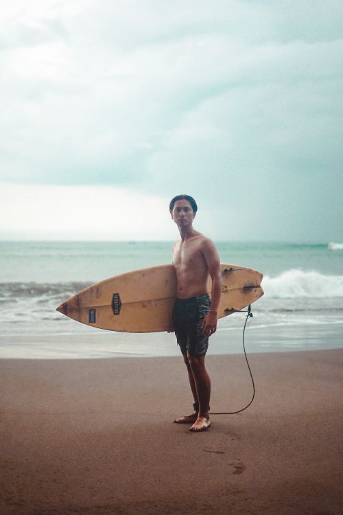 Free A surfer stands with his surfboard on a sandy beach, ready to hit the waves. Stock Photo