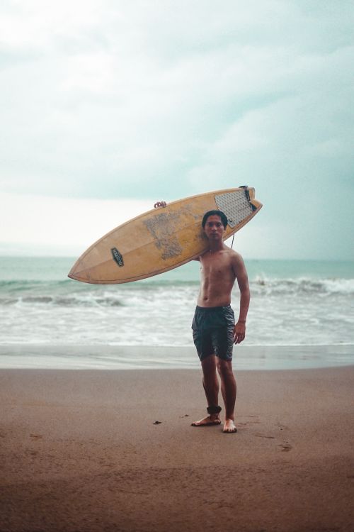 Free Shirtless man holding a surfboard on a sandy beach with cloudy skies and ocean waves. Stock Photo