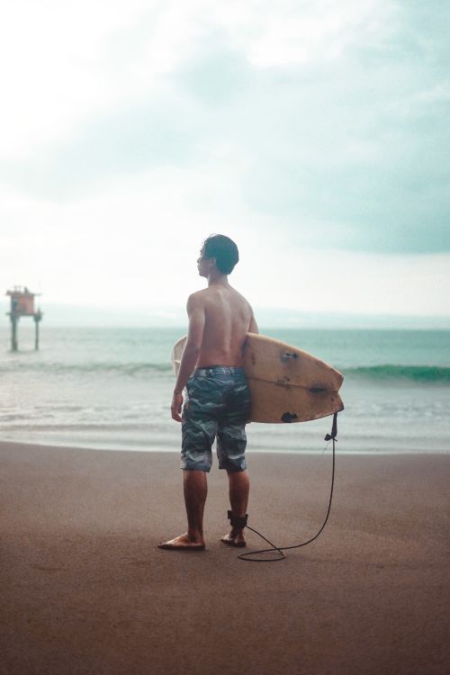 Free A surfer stands on a sandy beach holding a surfboard, looking at the ocean. Stock Photo