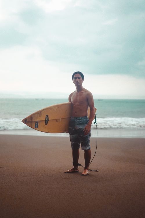 Free A surfer stands on a sandy beach with a surfboard, looking at the ocean under a cloudy sky. Stock Photo