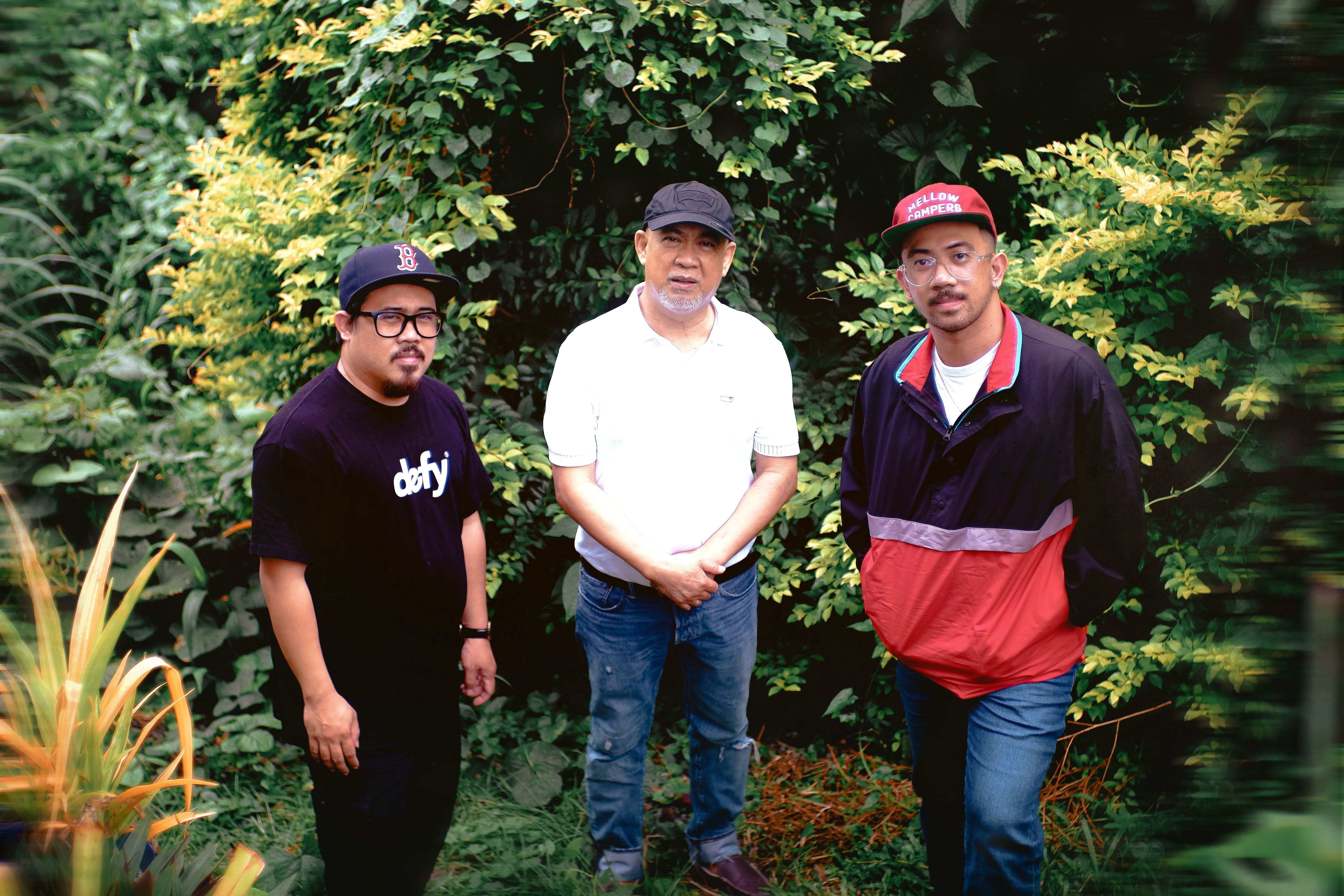 Three men posing outdoors surrounded by lush greenery, wearing casual clothing and caps.
