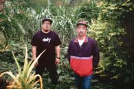 Two fashionable men in a lush garden wearing trendy streetwear, showcasing urban style.