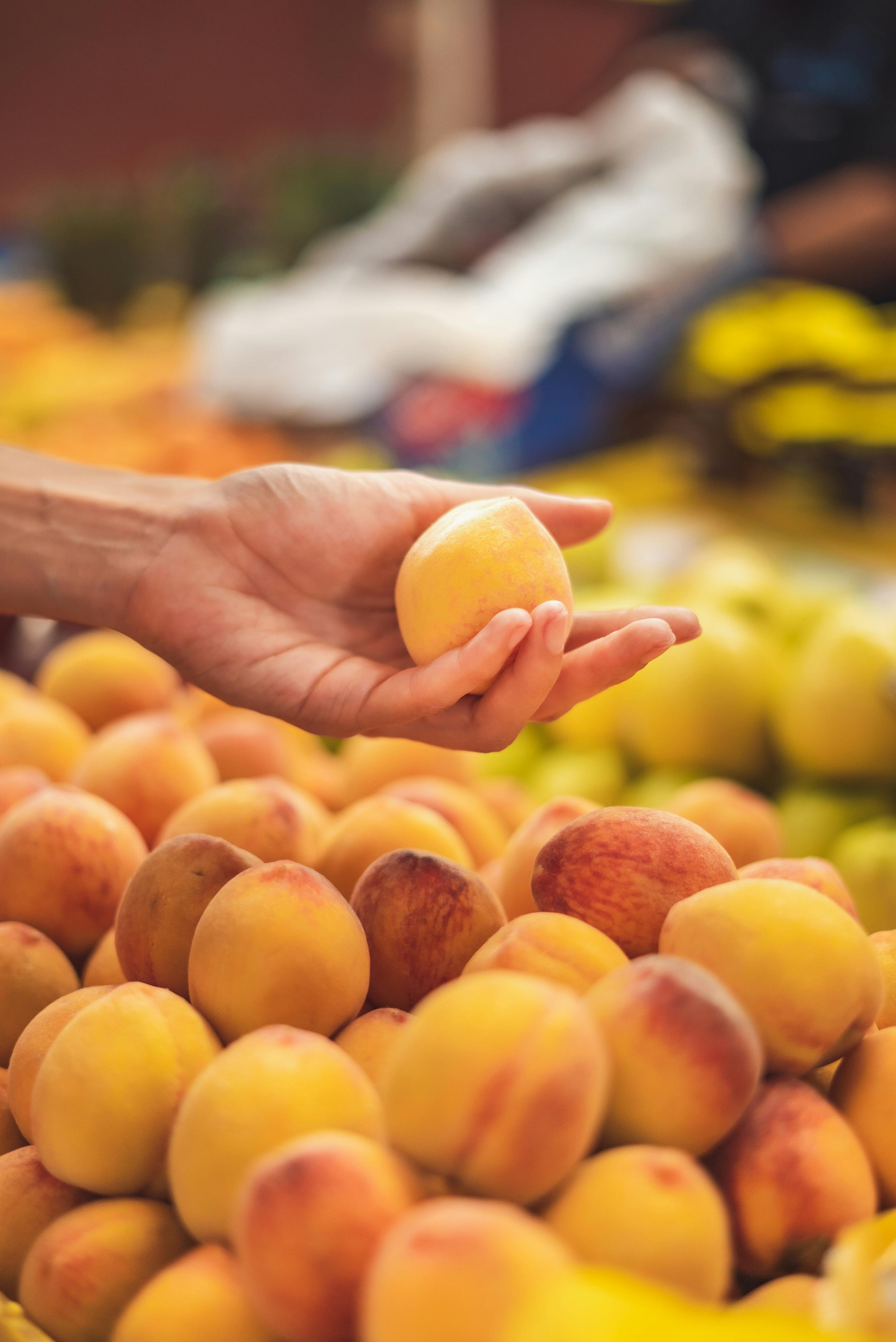 Close-up of a hand selecting a ripe peach in a vibrant market setting.