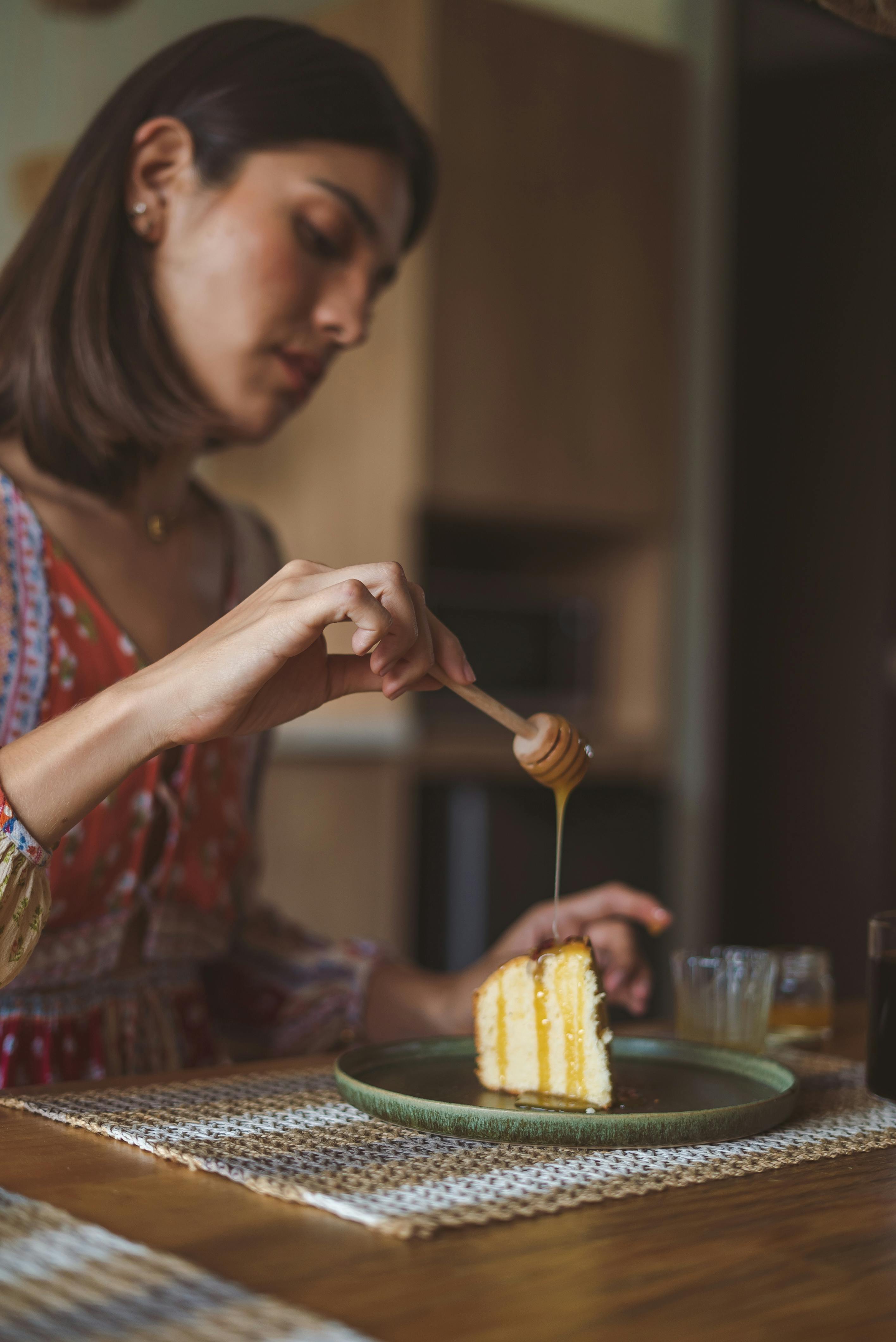 A woman drizzles honey over a layered cake slice on a plate, creating a cozy and inviting atmosphere indoors.