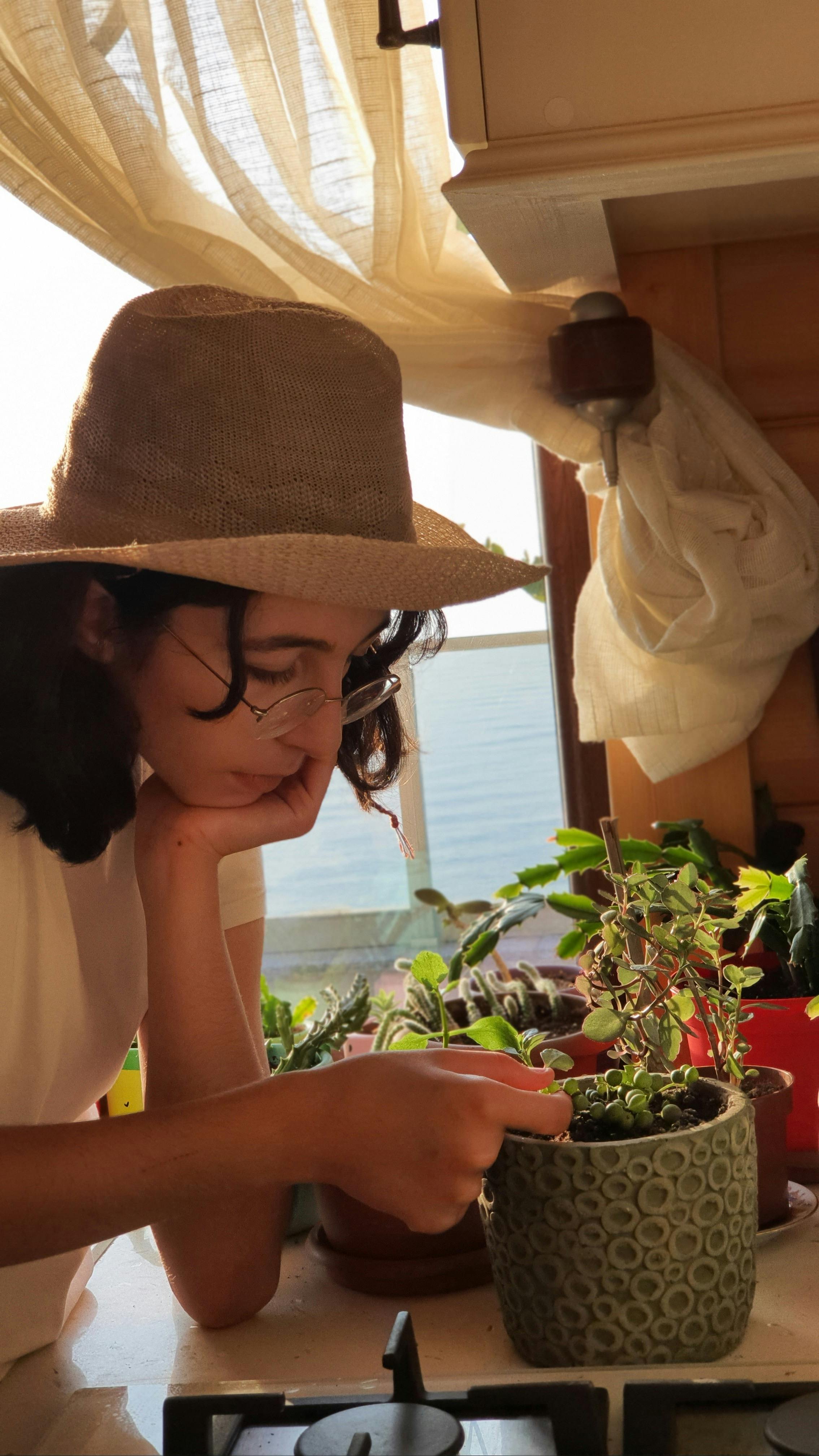 A young woman in a hat tends to potted plants by a window in a warm, sunlit kitchen setting.