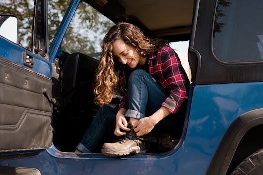 A woman in a flannel shirt smiling while tying her hiking boots in a blue off-road vehicle.