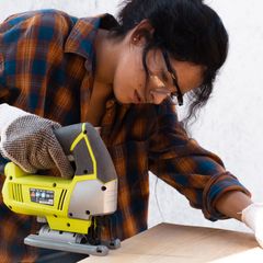 Woman in safety glasses and a checked shirt cutting a board with a yellow and grey compact jigsaw