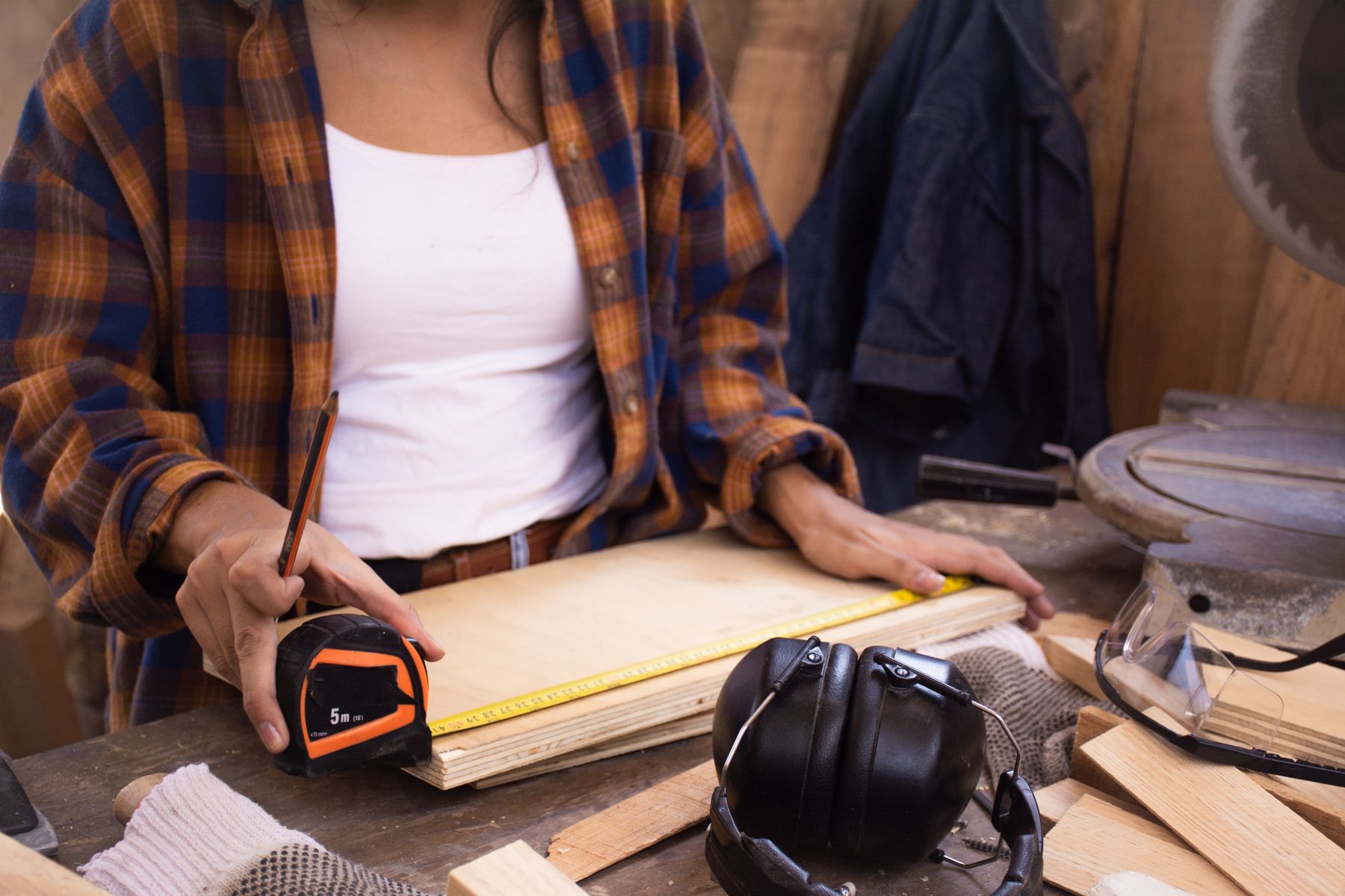 Carpenter measuring a wood panel with a tape measure beside protective hearing gear and workshop tools