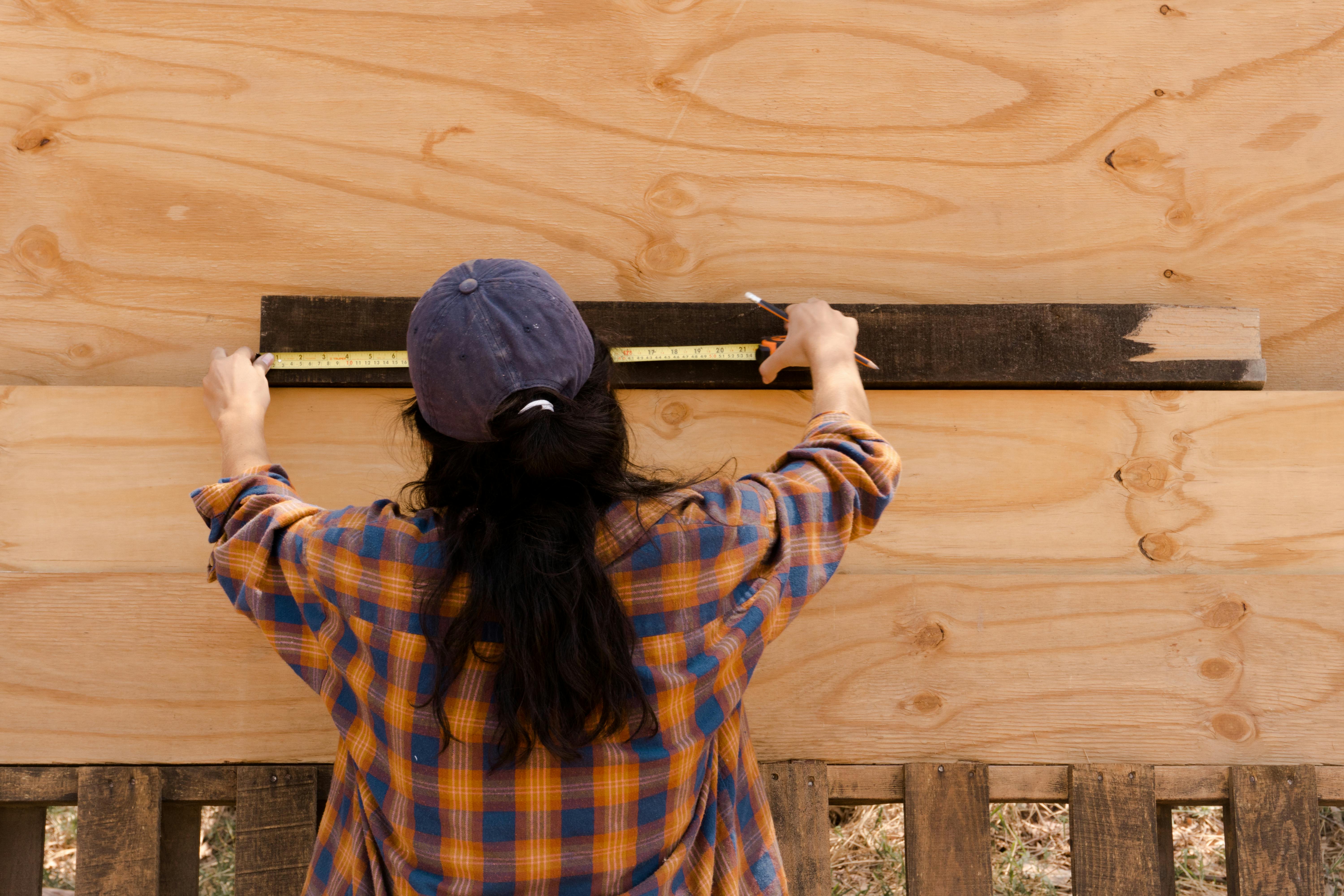 Person measuring a wooden plank with a tape measure. Back view wearing a blue cap and plaid shirt.
