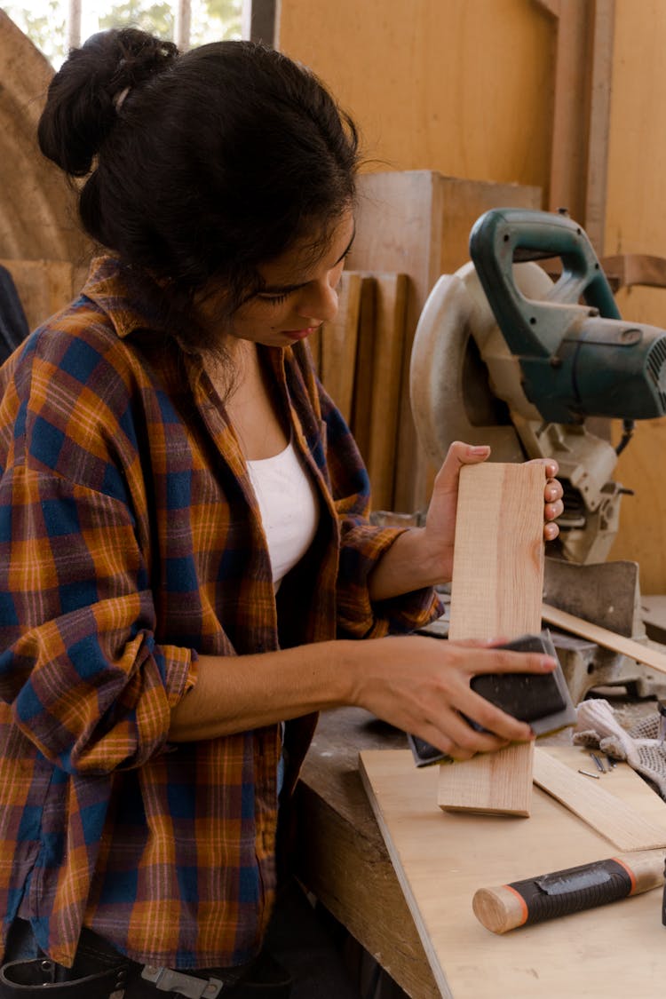 Woman In Orange And Blue Plaid Dress Shirt Sanding A Piece Of Wood