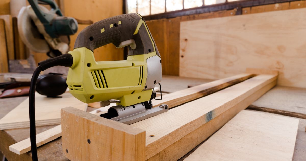 Yellow and gray corded jigsaw resting on a pine batten in a sunlit workshop, plywood sheets behind