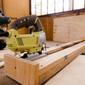 Yellow and grey corded jigsaw resting on a pine batten in a sunlit workshop, plywood sheets behind