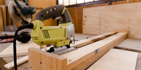 Yellow and gray corded jigsaw resting on a pine batten in a sunlit workshop, plywood sheets behind