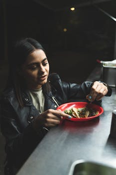 A woman in a leather jacket eating tacos at a food stand during nighttime.