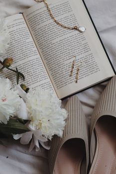 Chic still life featuring an open book, delicate flowers, and beige shoes on a bed.