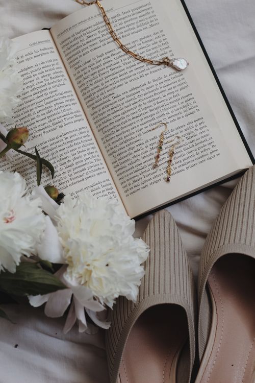 Free Chic still life featuring an open book, delicate flowers, and beige shoes on a bed. Stock Photo