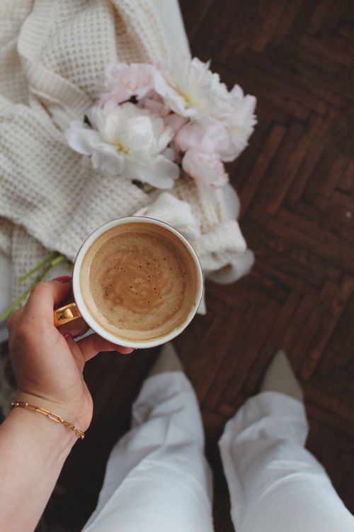 Free A hand holds a coffee cup above flowers and knit blanket, capturing a cozy mood. Stock Photo