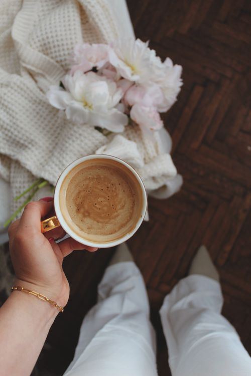 Free Cup of coffee held above soft blanket and flowers, creating a cozy morning vibe. Stock Photo