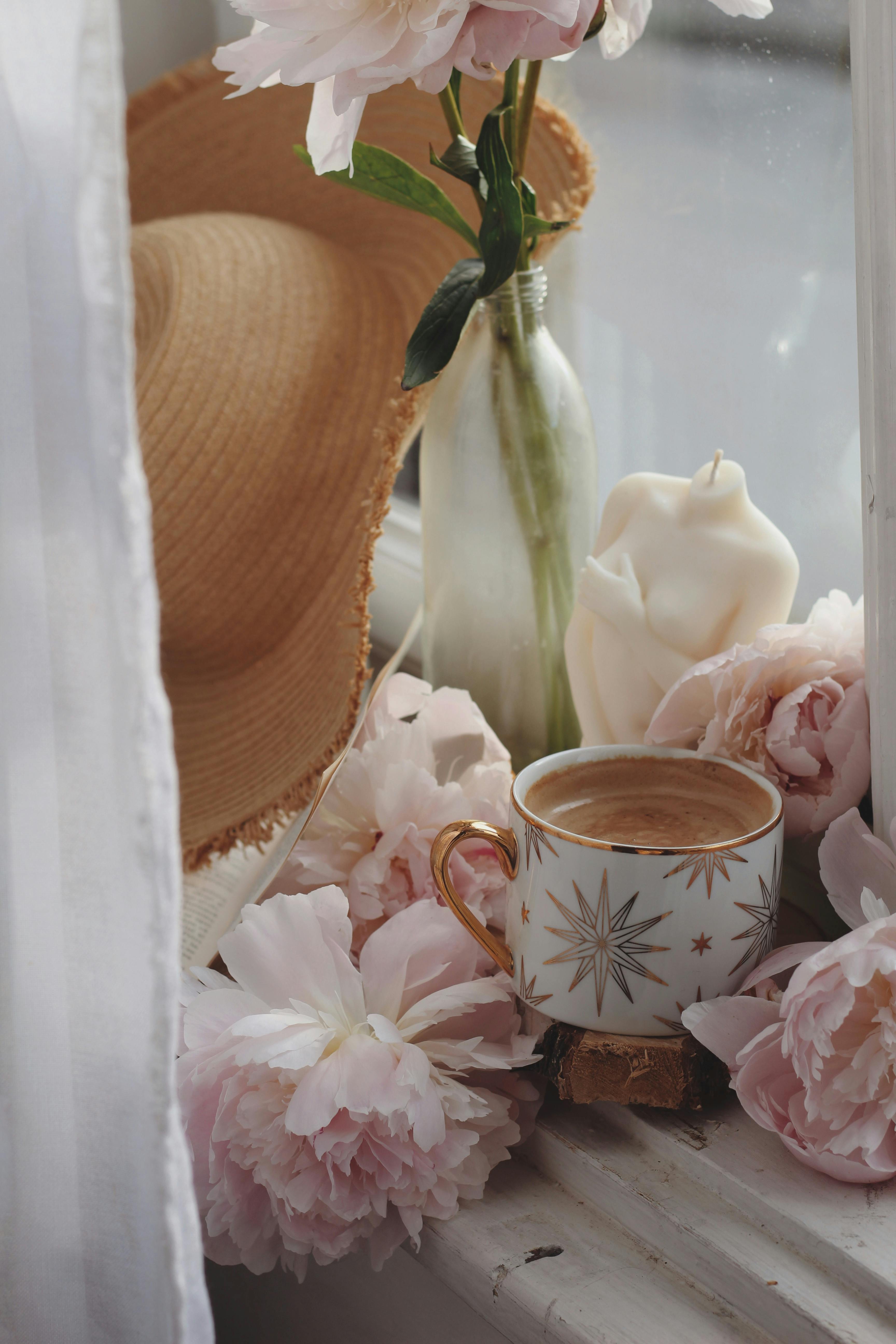 Free Elegant scene of a coffee cup with pink flowers, candle, and hat on a sunlit window ledge. Stock Photo