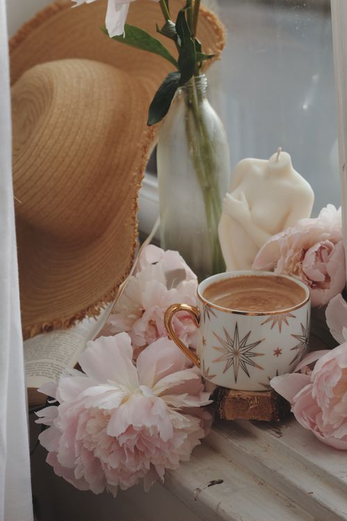 Free A serene arrangement featuring a coffee cup amidst pink flowers and a beach hat on a windowsill. Stock Photo
