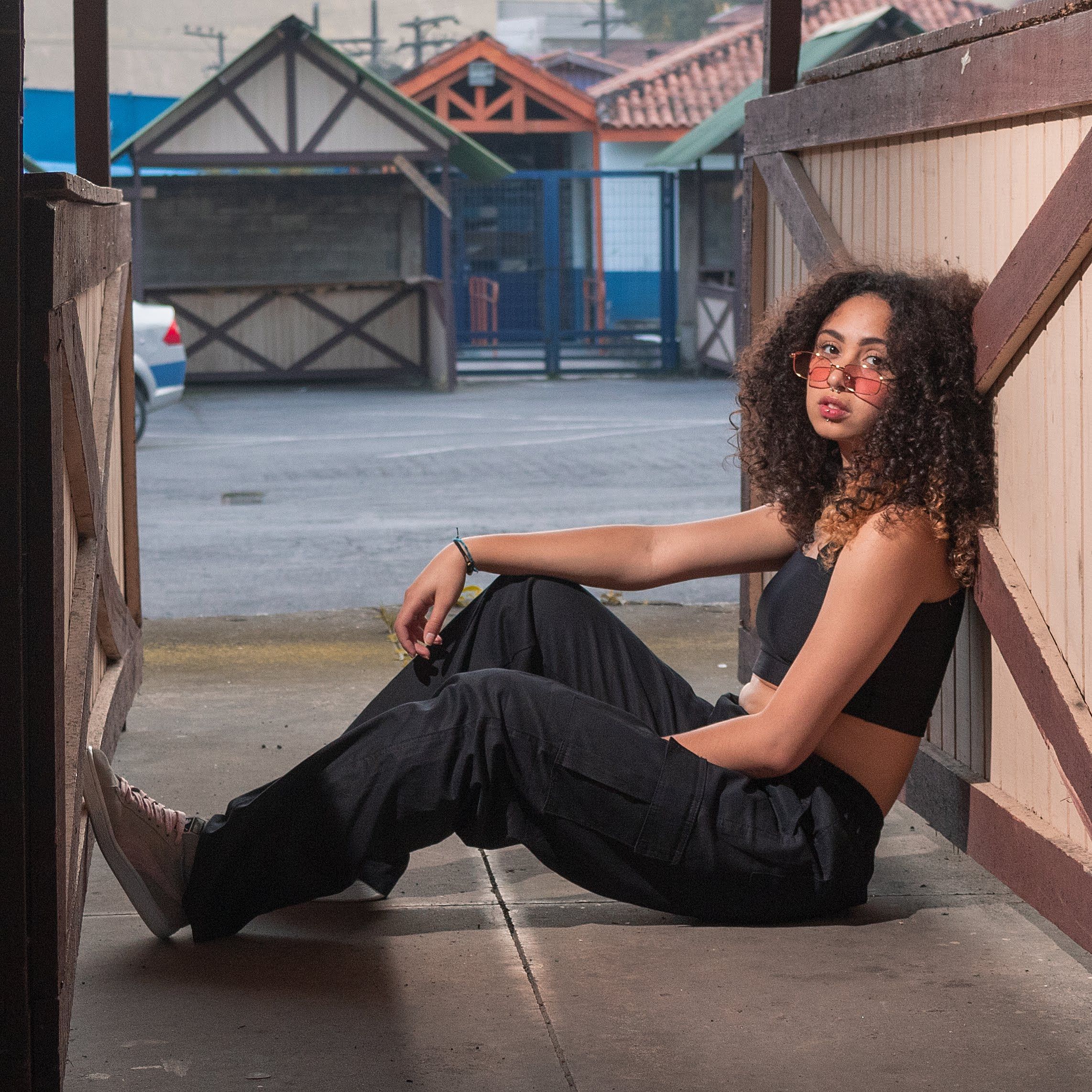 Young woman with curly hair in black outfit, sitting and posing in an urban outdoor setting.
