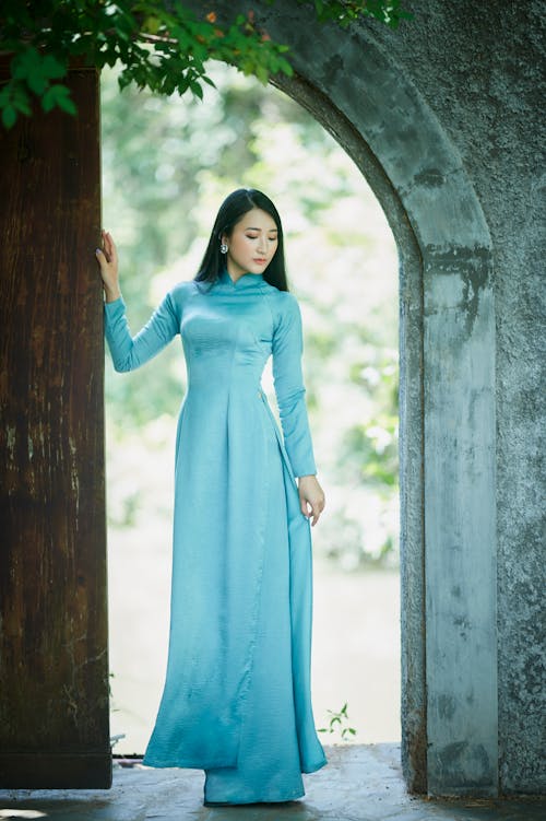 Woman in Blue Long Sleeve Dress Standing Beside Brown Wooden Door