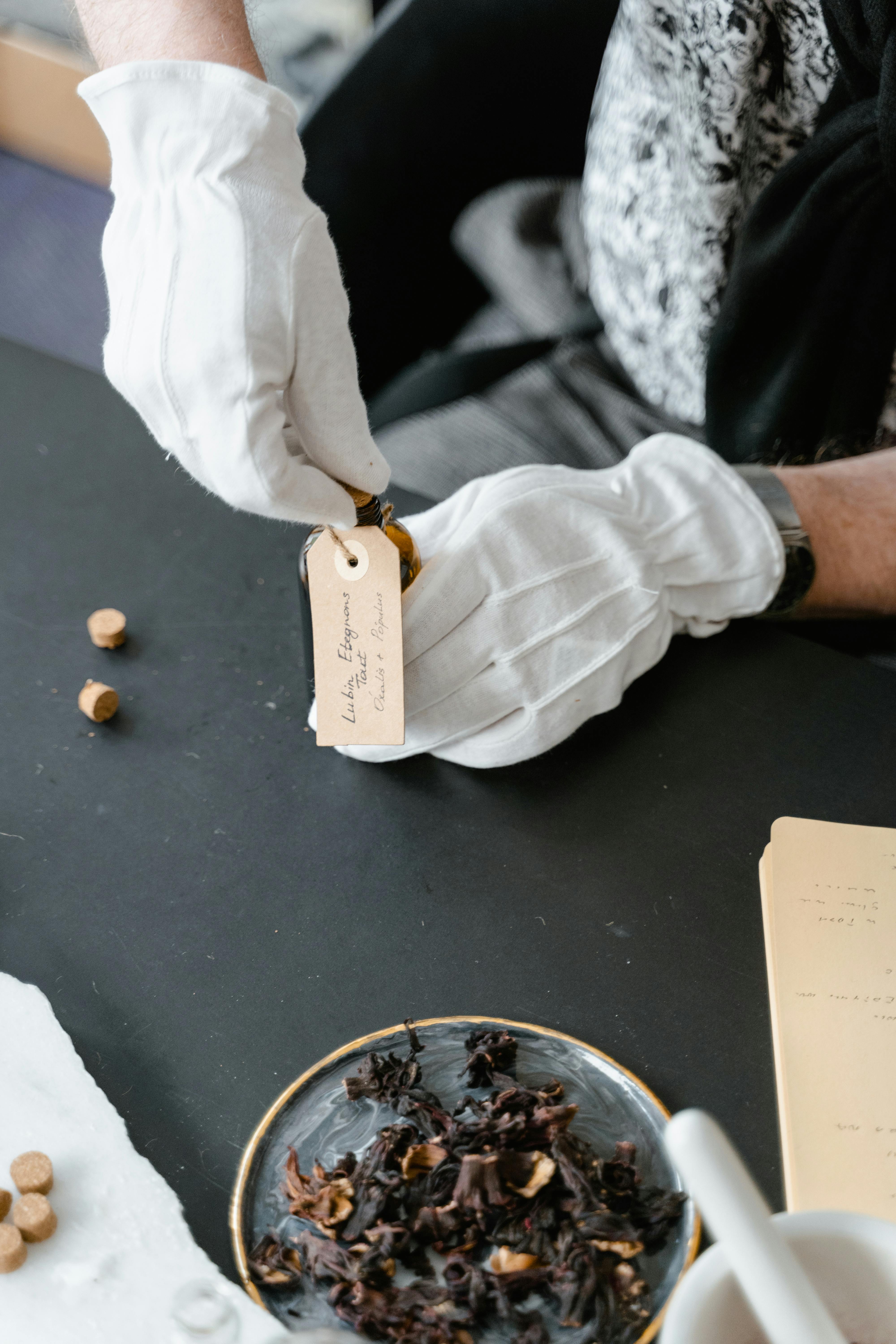 White-gloved hands carefully tagging a perfume bottle with dried ingredients nearby.