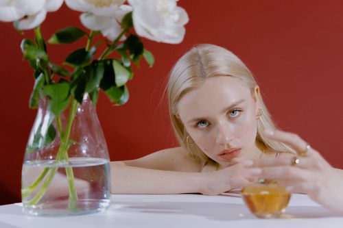 Free Portrait of a woman holding a perfume bottle beside a floral vase against a red background. Stock Photo
