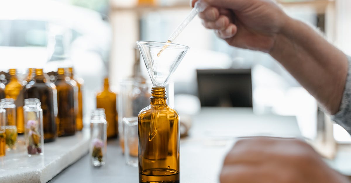 Hand pouring liquid into amber bottle using funnel, surrounded by glass jars indoors. Hand pouring liquid into amber bottle using funnel, surrounded by glass jars indoors.