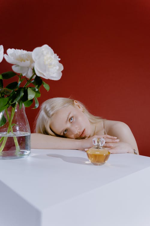 Free Blonde Girl Leaning on White Table With Bottle of Perfume and Glass Vase Stock Photo