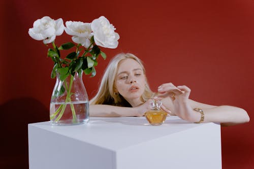 Woman with Blonde Hair Sitting Behind a Table with Vase and Bottle of Perfume