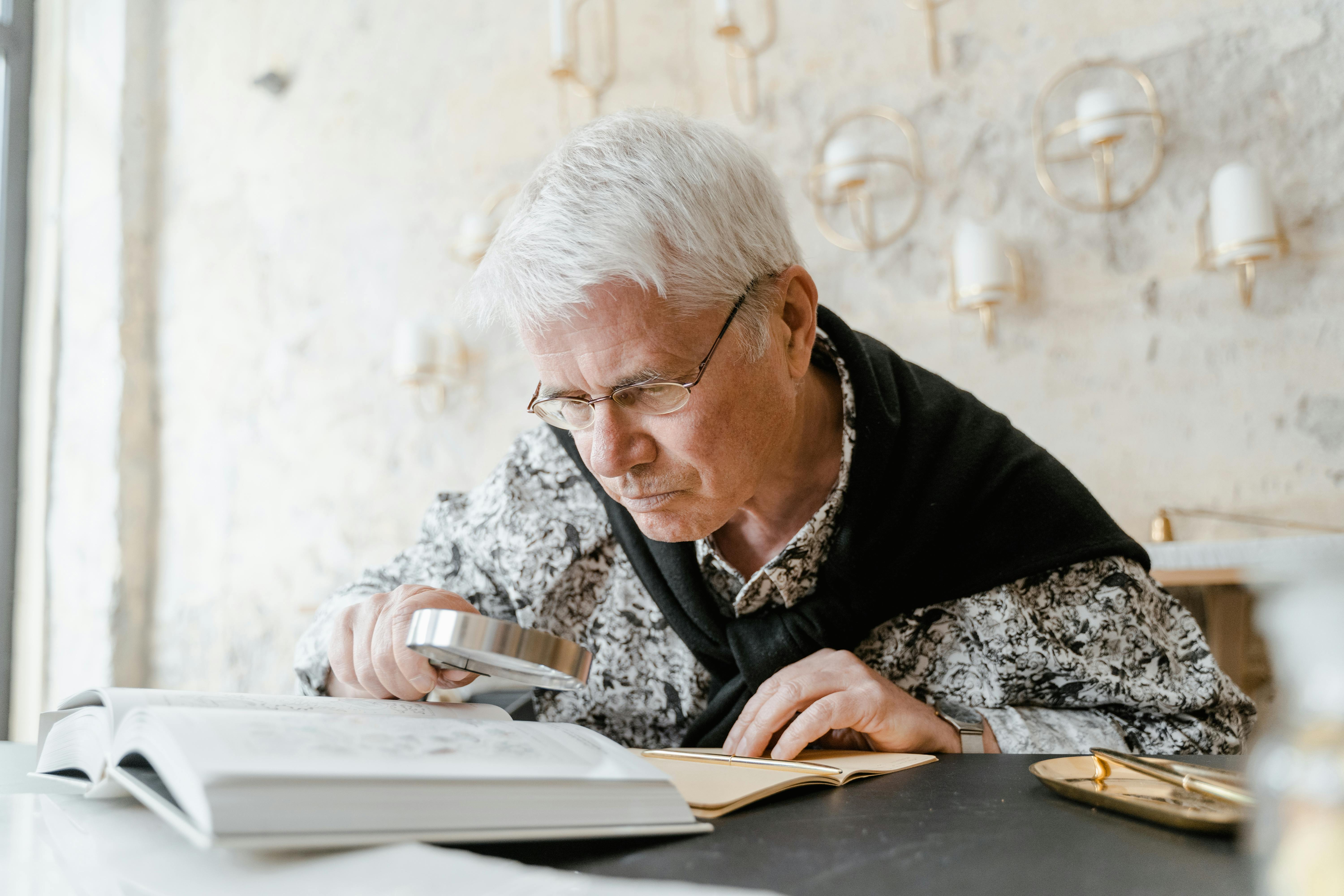 Elderly man using a magnifying glass to read books indoors at a cozy setting.