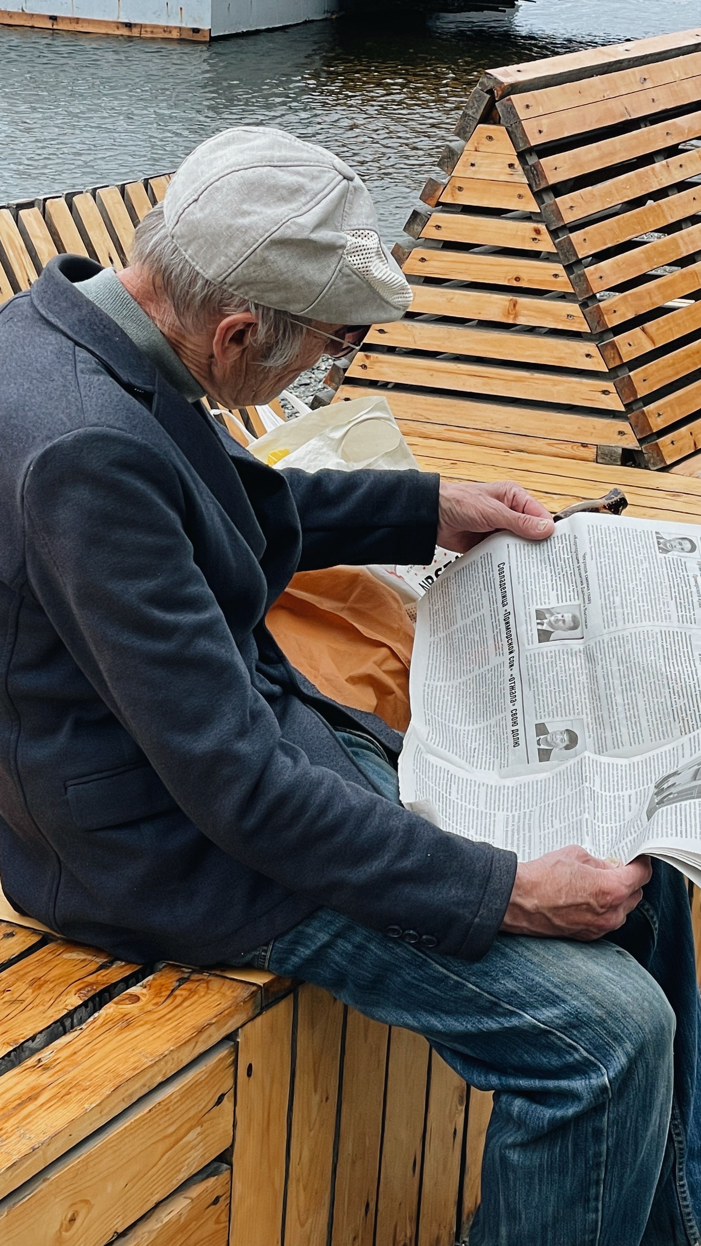 Senior man in a cap reading a newspaper outdoors on a uniquely designed wooden bench.