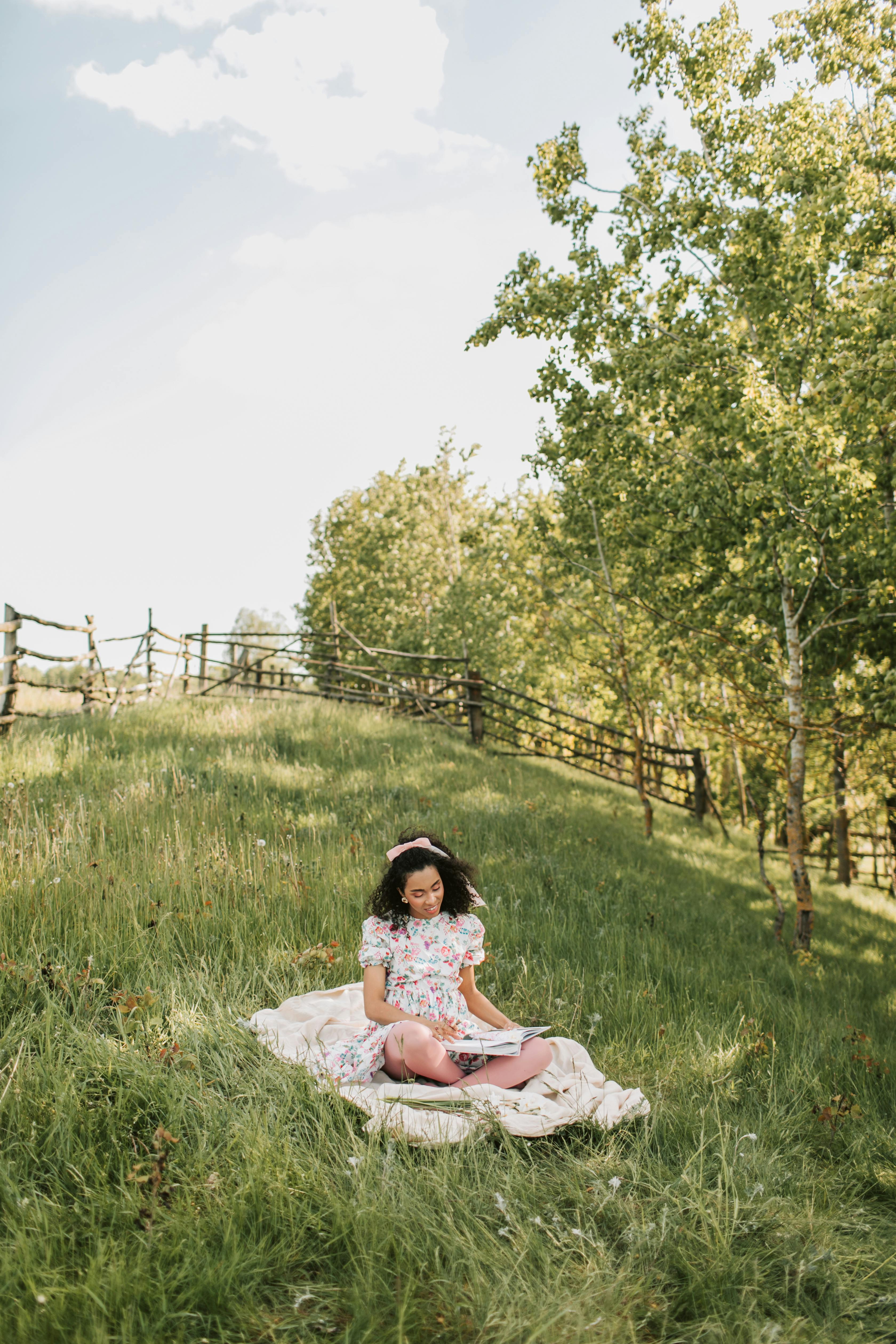 A young girl sits on a blanket in a sunny park, immersed in a book.