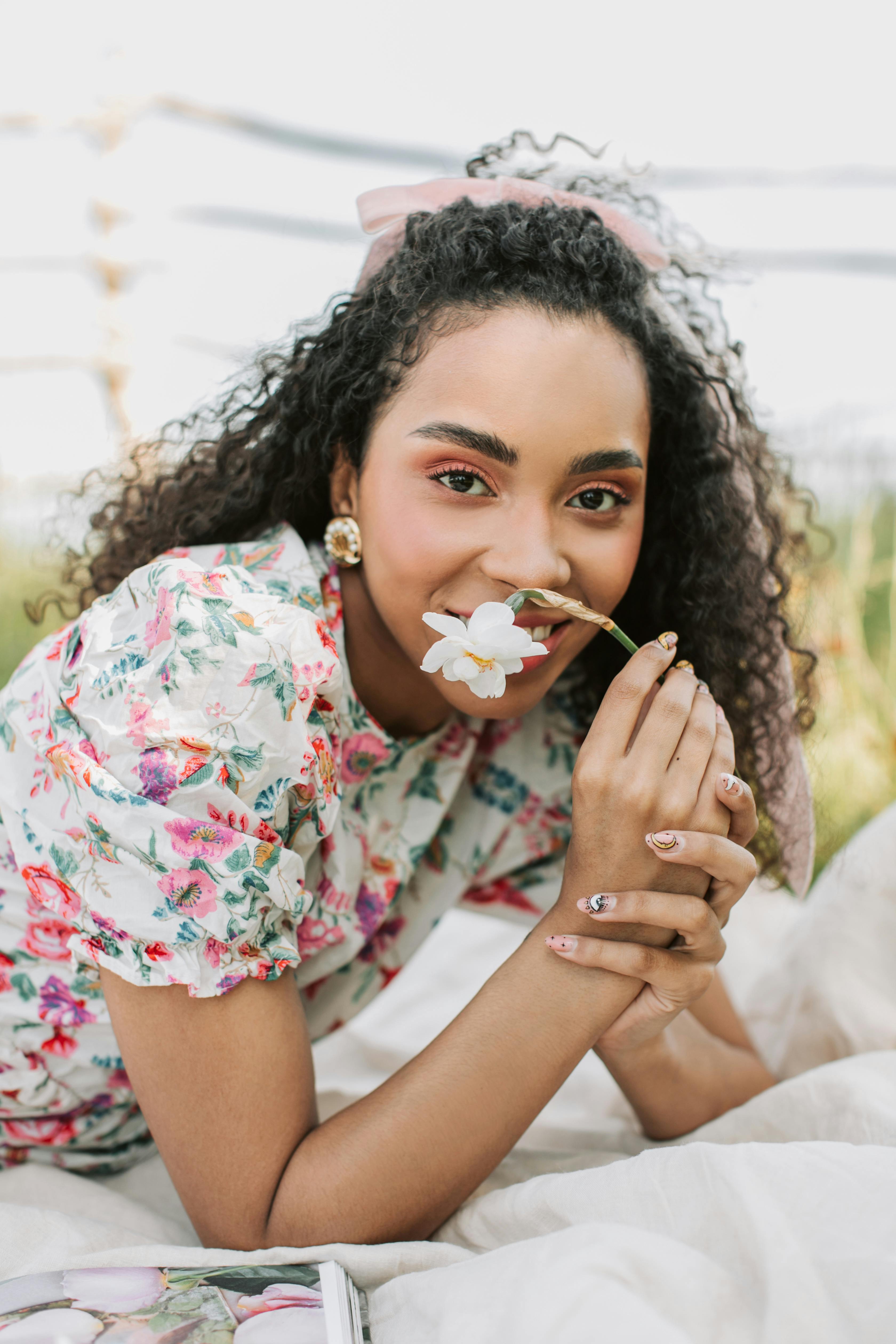 Cheerful woman holding a flower, wearing a floral dress, in an outdoor setting.