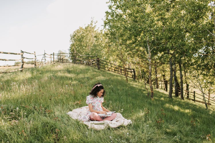 A Woman Wearing Dress Sitting On The Grass While Reading A Book