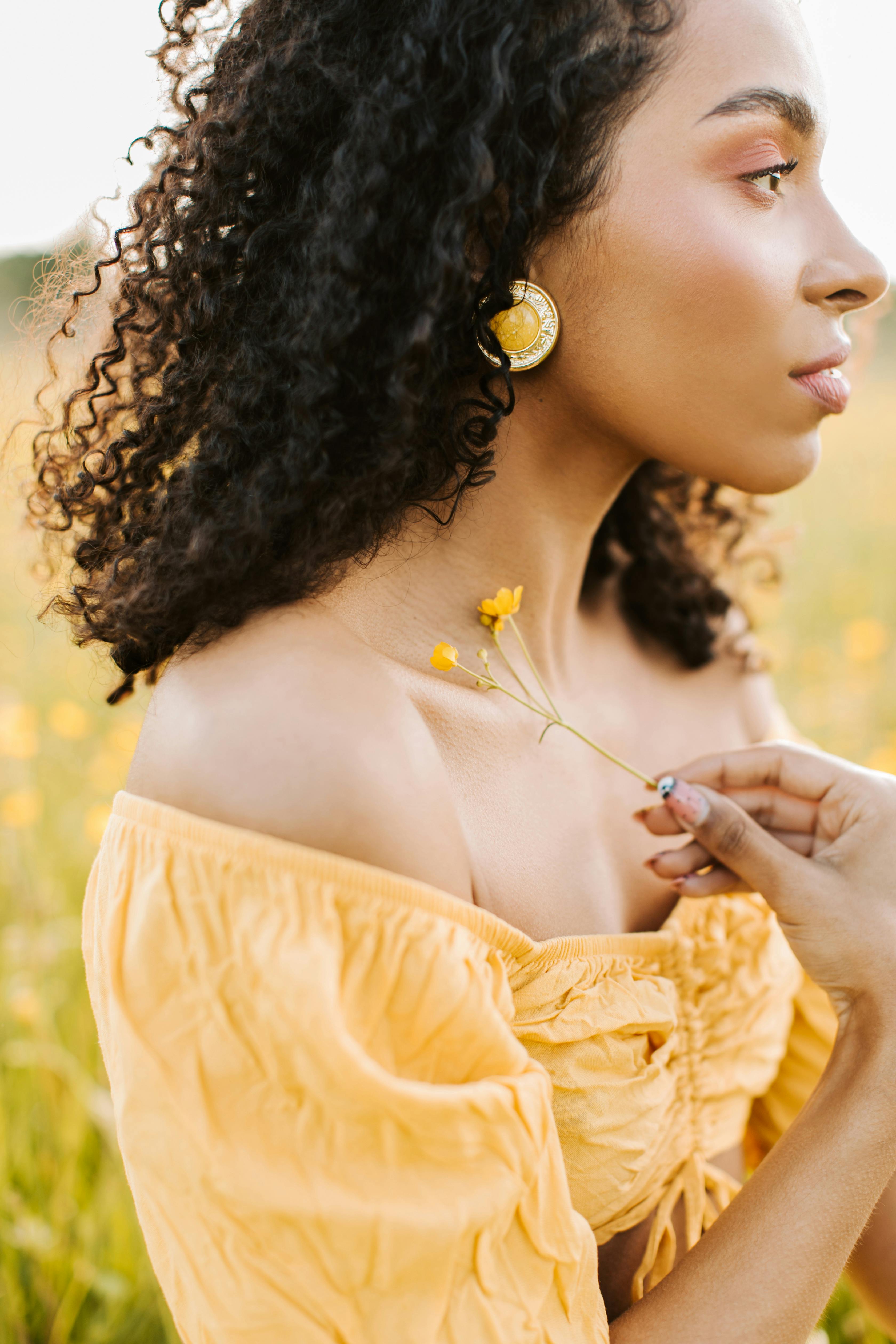 Side profile of a beautiful woman with curly hair and yellow dress holding a flower in a meadow.