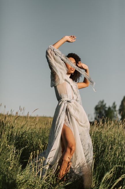 A woman poses gracefully in a sheer, flowing dress in a sunlit grassy field.